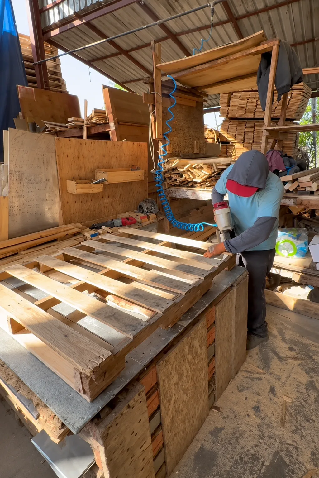 Worker driving nails into pallet deckboards with a pneumatic coil nailer at a timber workbench