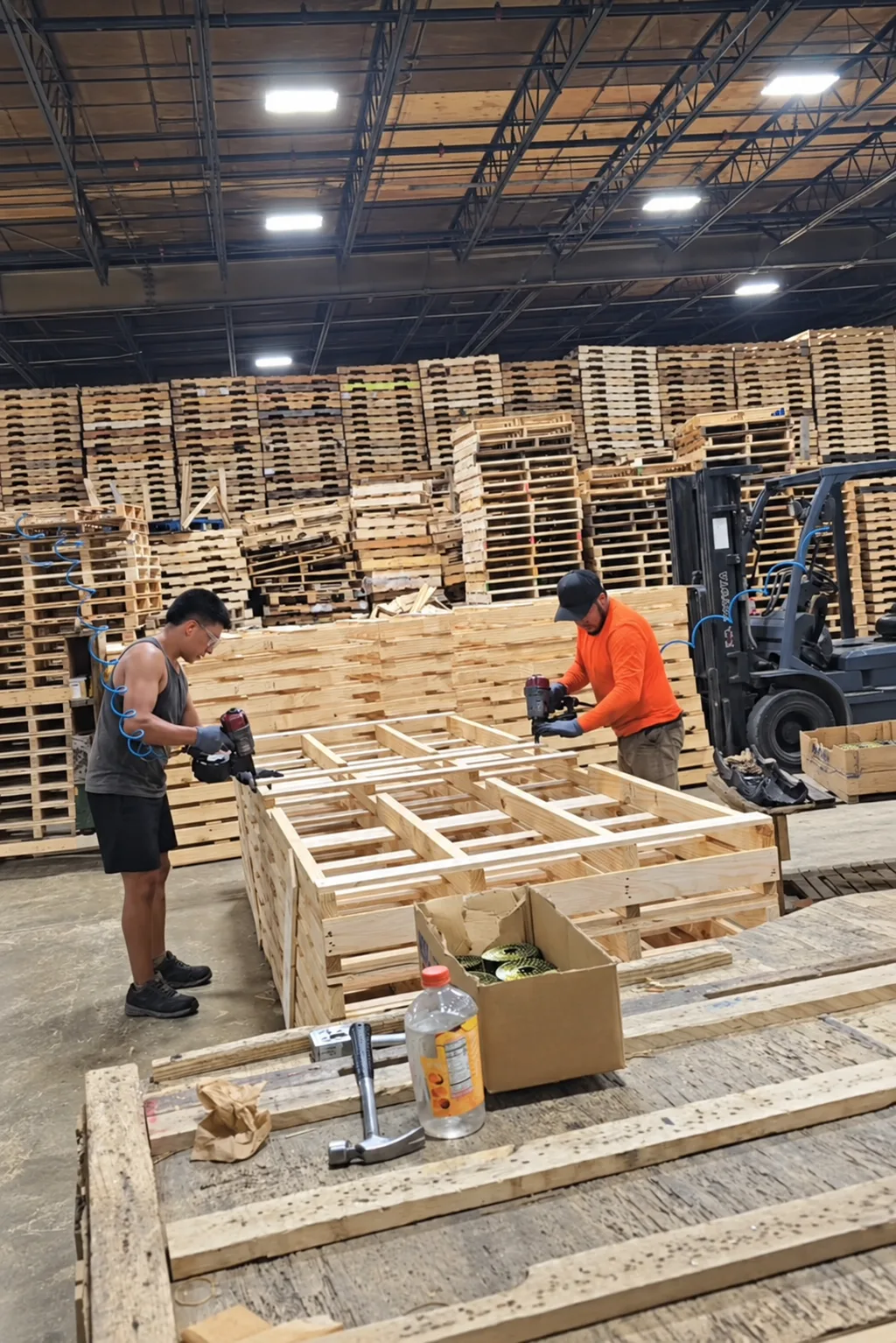 Two workers using coil nailers to assemble wooden pallets, with an open carton of nail coils in the foreground